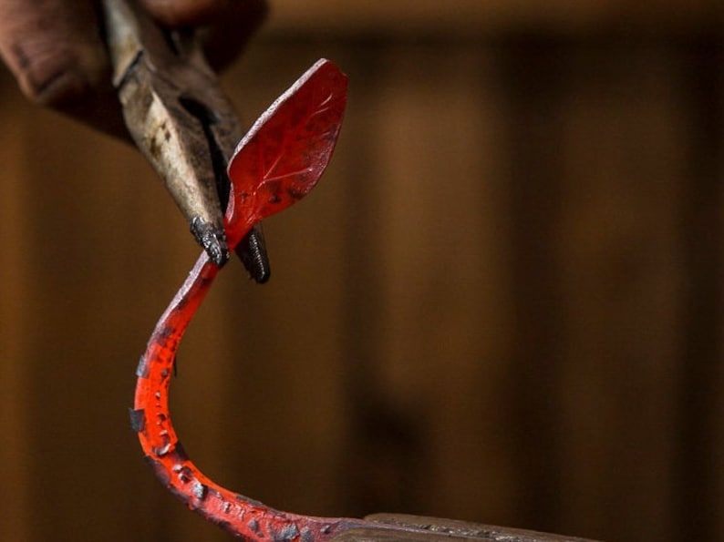 May include: A pair of pliers holds a red-hot metal leaf, which is being shaped into a curved form.