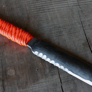 May include: A handmade knife with a black blade and a serrated edge. The handle is wrapped in bright orange cord. The knife rests on a weathered wooden surface, showcasing its rustic aesthetic. The blade measures approximately 10 inches in length.