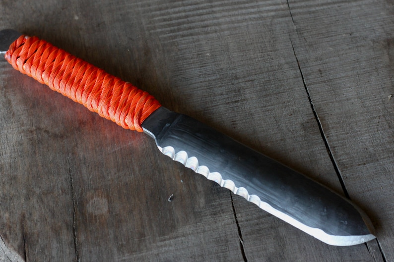 May include: A handmade knife with a black blade and a serrated edge. The handle is wrapped in bright orange cord. The knife rests on a weathered wooden surface, showcasing its rustic aesthetic. The blade measures approximately 10 inches in length.