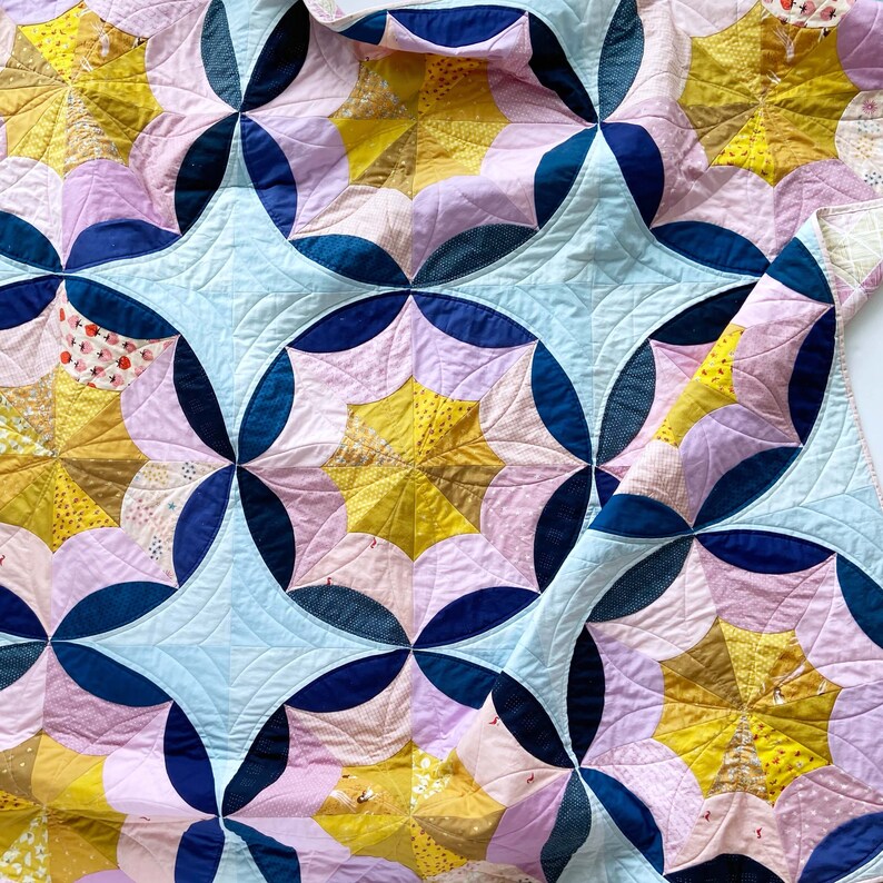 Puede incluir: Una colcha con un patr&oacute;n geom&eacute;trico de c&iacute;rculos superpuestos en tonos de azul, rosa y amarillo. La colcha est&aacute; hecha de retazos de tela y tiene un aspecto cosido a mano.