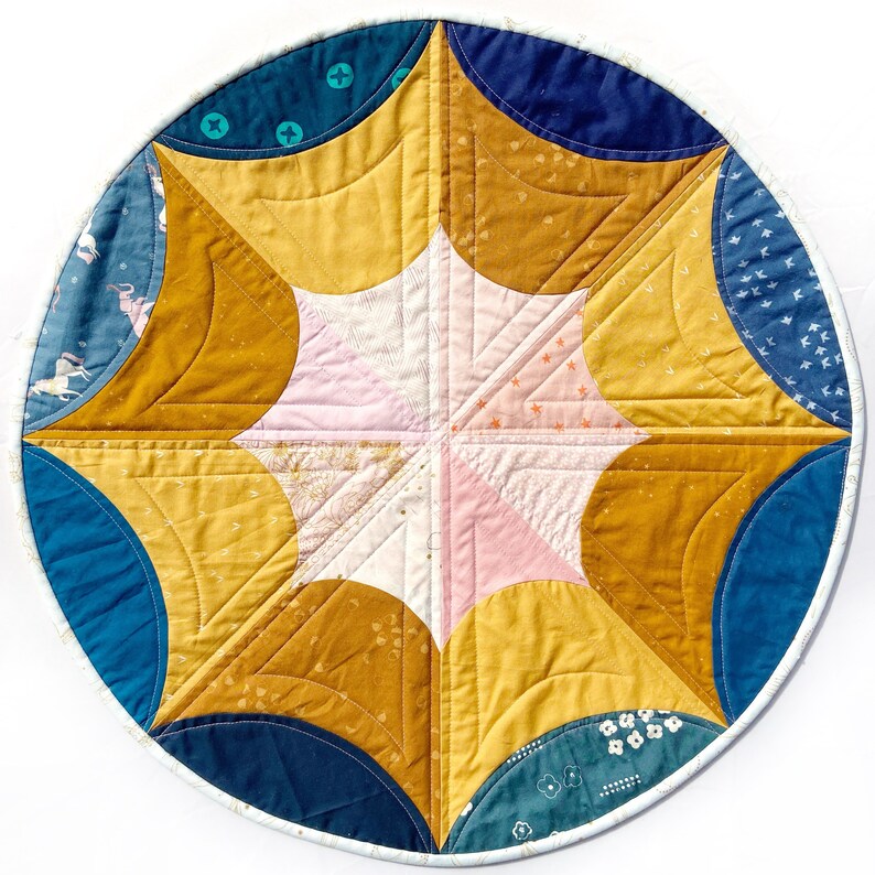 Puede incluir: Un tapiz de pared redondo acolchado con un dise&ntilde;o de estrella. El centro de la estrella es rosa y blanco, y los puntos exteriores son de color amarillo mostaza. El fondo es azul oscuro con acentos blancos.