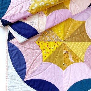 Puede incluir: Una colcha hecha de varias telas en tonos de rosa, amarillo, azul y blanco. La colcha est&aacute; hecha de formas curvas y est&aacute; doblada por la mitad. La colcha est&aacute; hecha con una variedad de patrones, incluyendo estrellas, flores y formas geom&eacute;tricas.