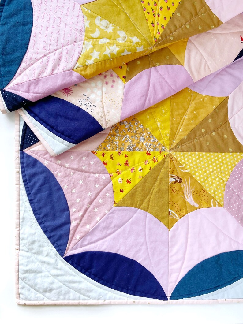 Puede incluir: Una colcha hecha de varias telas en tonos de rosa, amarillo, azul y blanco. La colcha est&aacute; hecha de formas curvas y est&aacute; doblada por la mitad. La colcha est&aacute; hecha con una variedad de patrones, incluyendo estrellas, flores y formas geom&eacute;tricas.