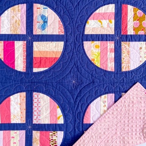 May include: A pink and white patchwork quilt with a navy blue backing. The quilt is made up of circles with a cross pattern in the centre. The quilt is quilted with a scalloped design.