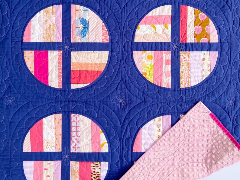 May include: A pink and white patchwork quilt with a navy blue backing. The quilt is made up of circles with a cross pattern in the centre. The quilt is quilted with a scalloped design.