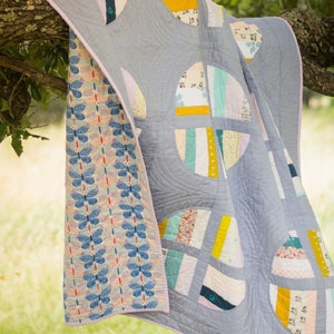 May include: A quilt with a patchwork design featuring circles, stripes, and floral patterns. The quilt is made of a variety of fabrics in shades of blue, pink, yellow, and green. The quilt is hanging from a tree branch.