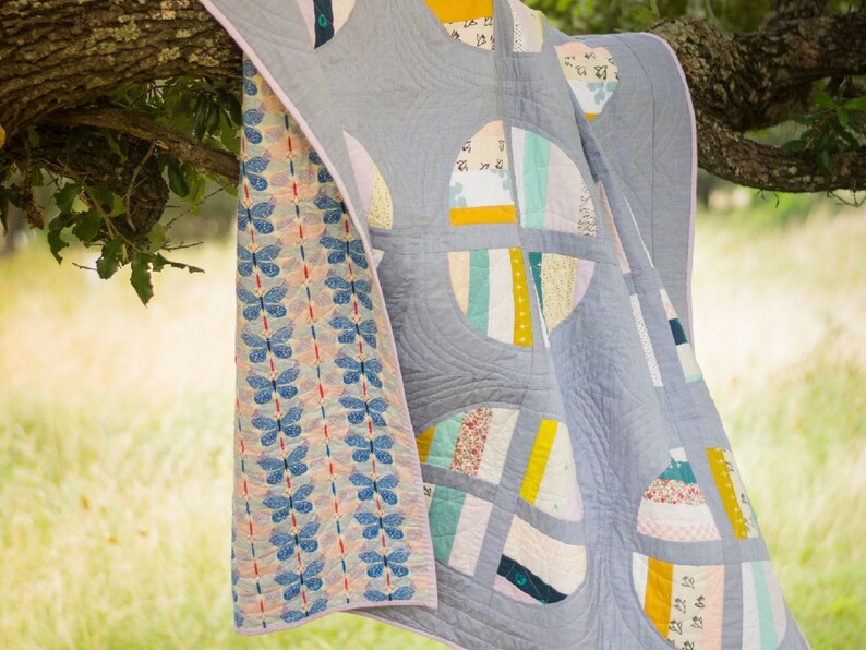 May include: A quilt with a patchwork design featuring circles, stripes, and floral patterns. The quilt is made of a variety of fabrics in shades of blue, pink, yellow, and green. The quilt is hanging from a tree branch.