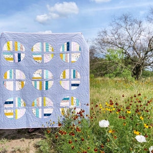 May include: A blue and white quilt with a geometric pattern of circles and squares. The quilt is hanging on a clothesline in a field of wildflowers.