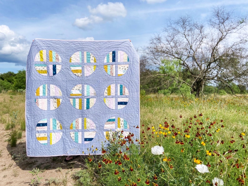 May include: A blue and white quilt with a geometric pattern of circles and squares. The quilt is hanging on a clothesline in a field of wildflowers.