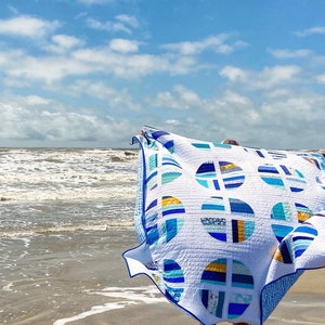 May include: A white quilt with blue and yellow geometric patterns, featuring circles and stripes. The quilt is being held up in the air on a beach with the sea in the background.