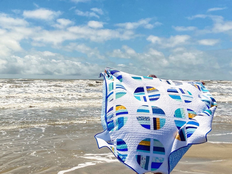 May include: A white quilt with blue and yellow geometric patterns, featuring circles and stripes. The quilt is being held up in the air on a beach with the sea in the background.