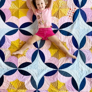 Puede incluir: Una ni&ntilde;a peque&ntilde;a con una camisa y pantalones cortos rosas yace sobre una colcha con un patr&oacute;n geom&eacute;trico de rosa, azul y amarillo. La colcha est&aacute; hecha de cuadrados de tela cosidos juntos para crear un patr&oacute;n repetitivo.