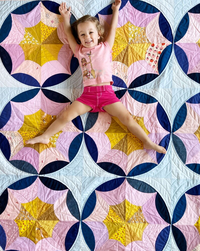 Puede incluir: Una ni&ntilde;a peque&ntilde;a con una camisa y pantalones cortos rosas yace sobre una colcha con un patr&oacute;n geom&eacute;trico de rosa, azul y amarillo. La colcha est&aacute; hecha de cuadrados de tela cosidos juntos para crear un patr&oacute;n repetitivo.