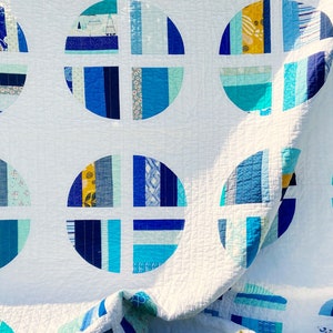 May include: A white quilt with a repeating pattern of blue and teal circles. The circles are made up of strips of fabric in various shades of blue and teal, and are arranged in a radial pattern. The quilt is folded in half, and the fabric is slightly wrinkled.
