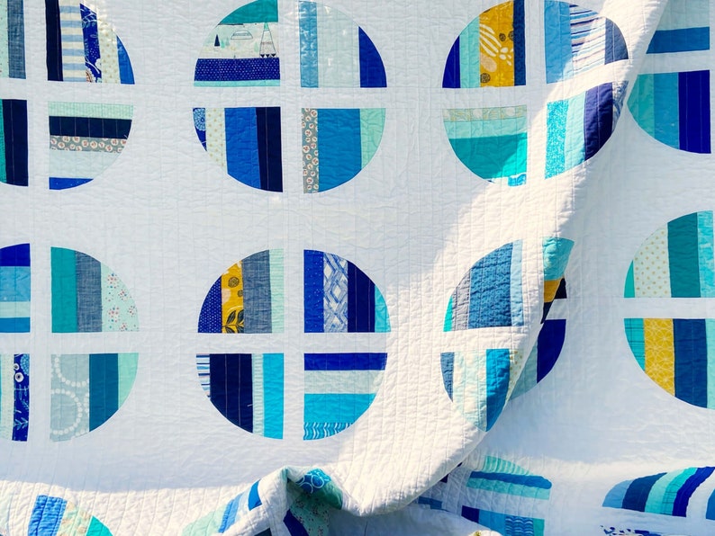 May include: A white quilt with a repeating pattern of blue and teal circles. The circles are made up of strips of fabric in various shades of blue and teal, and are arranged in a radial pattern. The quilt is folded in half, and the fabric is slightly wrinkled.