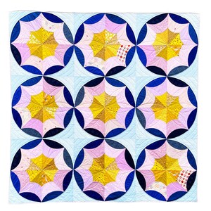 Puede incluir: Una colcha con fondo azul y blanco y nueve parches circulares. Cada parche es un patr&oacute;n de estrella rosa y amarilla.