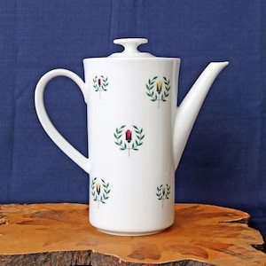 May include: White ceramic coffee pot with a lid and a curved handle. The pot is decorated with repeating floral designs in red, yellow, and green. The pot is on a wooden surface against a blue background.