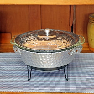 May include: A vintage silver chafing dish with a glass lid, set on a black metal stand. The dish has a hammered texture and decorative handles. The lid features a floral design.