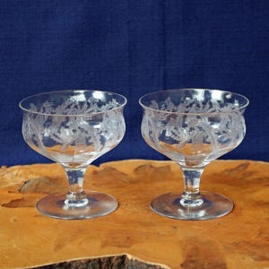 May include: Two clear, stemmed glasses with etched floral designs. The glasses have a rounded bowl shape and sit on a circular base. The background is a dark blue color, and the glasses are on a wooden surface.