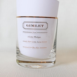 May include: A clear glass with a gold rim and a white paper insert. The paper lists the ingredients for a Gimlet cocktail: ice, 2 lime wedges, 1 1/2 oz. lime juice, and 3 oz. dry gin. The word "Gimlet" is in a decorative font.