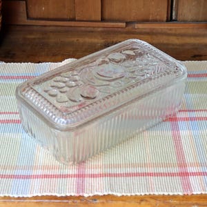 May include: A rectangular clear glass butter dish with a lid featuring a molded fruit design on top. The base and lid have vertical ribbed sides, and the lid is decorated with embossed grapes and leaves, providing a textured appearance.