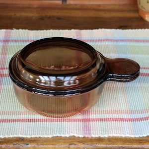 May include: A brown glass casserole dish with a lid and a handle. The dish is round with a slightly flared base. The lid fits snugly on top. The handle is attached to the side of the dish.