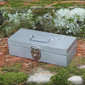 May include: A grey metal toolbox with a hinged lid and a silver latch. The toolbox has a metal handle on the top.