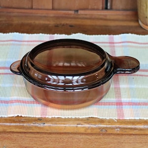 May include: A round, amber-colored glass casserole dish with a matching lid. The dish has two handles and is sitting on a plaid patterned placemat. The dish is made of a transparent material.