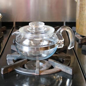 May include: A clear glass teapot with a lid and handle sits on a black stovetop. The teapot has a silver band around its middle. The burner is made of dark metal. A copper-colored pot is visible in the background.