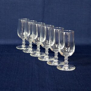 May include: A set of six clear glass cordial glasses arranged in a diagonal line. Each glass features a smooth, cylindrical bowl and a stem decorated with three stacked spherical glass beads, supported by a circular, flat glass base.