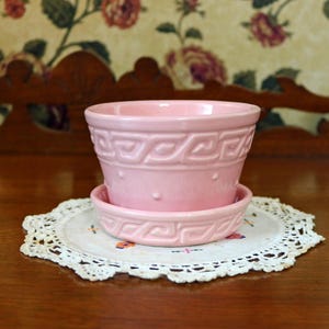 May include: A light pink ceramic flower pot with a matching saucer. The pot features a raised Greek key pattern around the upper rim and a series of small, evenly spaced raised dots encircling the middle section of the exterior surface.