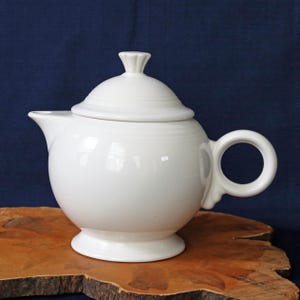 May include: A white ceramic teapot with a lid and a rounded body. The teapot has a handle and a spout. The lid has a small, decorative finial. The teapot is sitting on a wooden surface against a dark blue background.