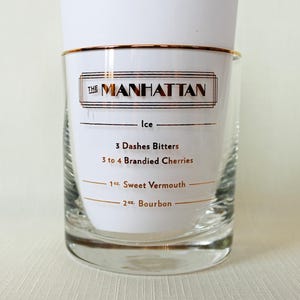 May include: A clear glass with a gold rim and the text "THE MANHATTAN" in gold. The glass has a white label with the recipe for a Manhattan cocktail, including bitters, cherries, sweet vermouth, and bourbon.