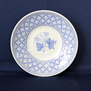 May include: A white ceramic saucer with a blue floral design. The saucer features a central image of blue leaves and berries, surrounded by a patterned border of blue geometric shapes and stars. The overall design is delicate and detailed.
