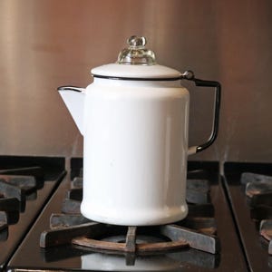 Peut inclure: Une cafetière en émail blanc avec une poignée et une garniture noires, posée sur une cuisinière noire. La cafetière a un bouton en verre transparent sur le couvercle. La cuisinière a quatre brûleurs.
