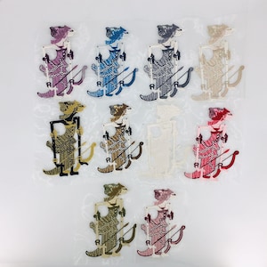 May include: A collection of ten embroidered patches featuring a stylized archer figure with a bow and arrow. The patches come in various colors, including purple, blue, gray, beige, black, brown, white, and red. Each patch has the letter "A" on the archer's foot.