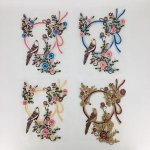 May include: Four embroidered patches featuring a floral wreath with two birds inside. The wreaths are in blue, pink, and gold with brown birds. The patches are on a white background.