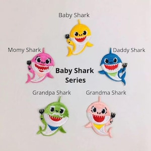 Może przedstawiać: Zestaw sześciu naszywek do prasowania z postaciami Baby Shark. Naszywki są żółte, różowe, niebieskie, zielone i jasnoróżowe. Każda naszywka przedstawia rekina z innym wyrażeniem twarzy i widelcem w płetwie. Tekst "Baby Shark Series" jest nadrukowany czarnym kolorem pod naszywkami.