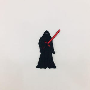 Op de afbeelding: Geborduurde patch met een zwart gekleed figuur met een rood lichtzwaard. De patch is een gedetailleerd, gestileerd ontwerp, perfect voor het toevoegen aan kleding of accessoires. Het figuur heeft een capuchon en het lichtzwaard is felrood.