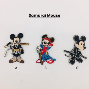 Könnte beinhalten: Drei gestickte Aufnäher mit einer Mausfigur in Samurai-Kleidung. Die Aufnäher sind mit A, B und C beschriftet. Aufnäher A ist schwarz und beige, B ist rot und blau und C ist schwarz. Der Text "Samurai Mouse" steht über den Aufnähern.