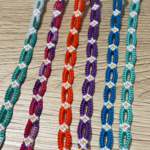 May include: A collection of handmade friendship bracelets in various colors. Each bracelet features a repeating pattern of woven knots in shades of teal, pink, orange, purple, and blue, accented with white and yellow details. The bracelets are displayed on a light wood surface.