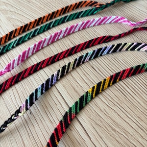 May include: A collection of colorful friendship bracelets with diagonal striped patterns. The bracelets feature various color combinations, including orange and black, green and black, pink and white, and red and black. The bracelets are displayed on a wooden surface.