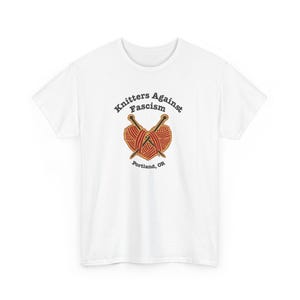 Könnte beinhalten: Ein weißes kurzärmeliges T-Shirt mit einer Grafik aus zwei gekreuzten Stricknadeln und einem herzförmigen roten Wollknäuel. Der Text "Knitters Against Fascism" ist darüber gebogen, mit "Portland, OR" in schwarzer Schrift darunter.