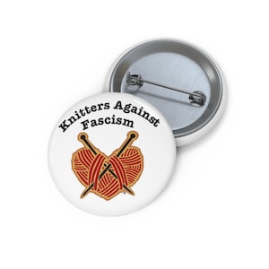Könnte beinhalten: Ein runder Ansteckbutton mit weißem Hintergrund und dem schwarzen Schriftzug Knitters Against Fascism über einer Grafik aus zwei gekreuzten Stricknadeln und einem herzförmigen roten Wollknäuel. Die Rückseite zeigt eine silberne Metallnadel.