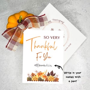 以下が含まれることがあります： オレンジ色の文字で「SO VERY Thankful For You」と書かれた白いカード。カードには「Brenda & Liz」と書かれたハートと、秋の色の花柄のデザインが施されています。カードは、小さなオレンジ色のカボチャと格子縞のリボンが置かれたテーブルの上にあります。