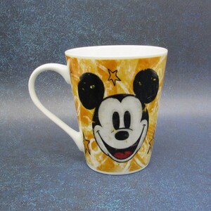Puede incluir: Una taza de caf&eacute; de cer&aacute;mica amarilla con una imagen de dibujos animados de Mickey Mouse. La taza tiene un asa blanca y un contorno negro alrededor de la imagen.