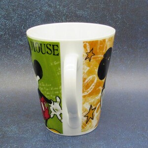 Puede incluir: Una taza de cer&aacute;mica blanca con un dise&ntilde;o verde, amarillo y negro con Mickey Mouse. La taza tiene las palabras "Mickey Mouse" impresas en el lateral.