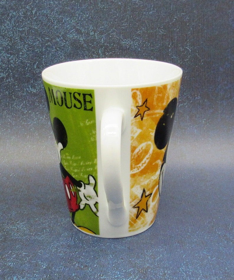 Puede incluir: Una taza de cer&aacute;mica blanca con un dise&ntilde;o verde, amarillo y negro con Mickey Mouse. La taza tiene las palabras "Mickey Mouse" impresas en el lateral.