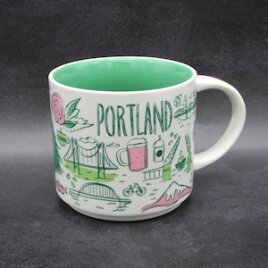 May include: A white ceramic mug with a mint green interior featuring a green and pink illustrated design of Portland landmarks, including bridges, a bicycle, a rose, and a beer mug, with the word PORTLAND printed in large green letters.