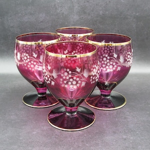 May include: A set of four deep magenta colored glass goblets featuring etched white grape vine patterns and gold rimmed edges. Each goblet has a rounded bowl, a short stem, and a wide circular base, all crafted from translucent glass.